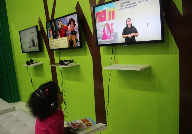Uma menina está sentada e segura um livro em seu colo. Ela usa fones de ouvido e olha para uma tela onde é exibido um livro audiovisual em múltiplos formatos acessíveis