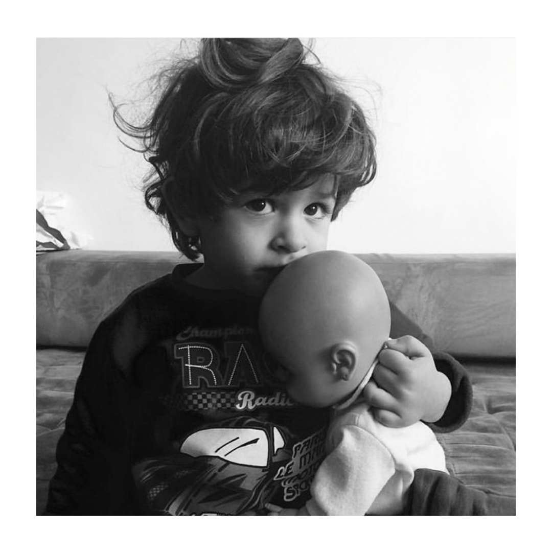 Foto em preto e branco de um menininho segurando uma boneca bebê. Ele encosta a boca na cabeça da boneca e olha de forma meiga para a câmera.