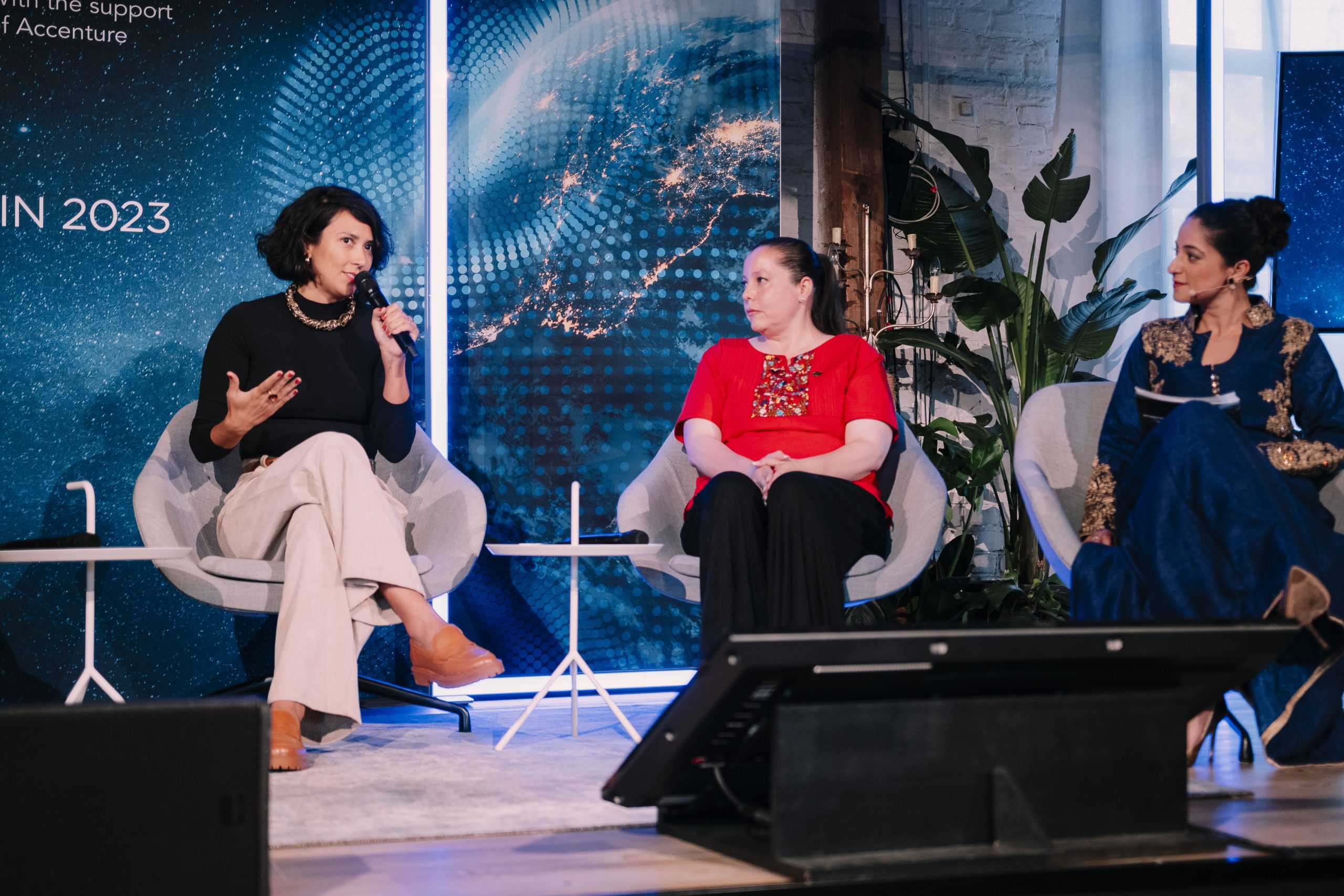 A foto mostra Thais Pereira Martins, Coordenadora de Advocacy da Mais Diferenças, sentada ao lado de duas mulheres, falando ao microfone, na cerimônia do Intercultural Innovation Hub.