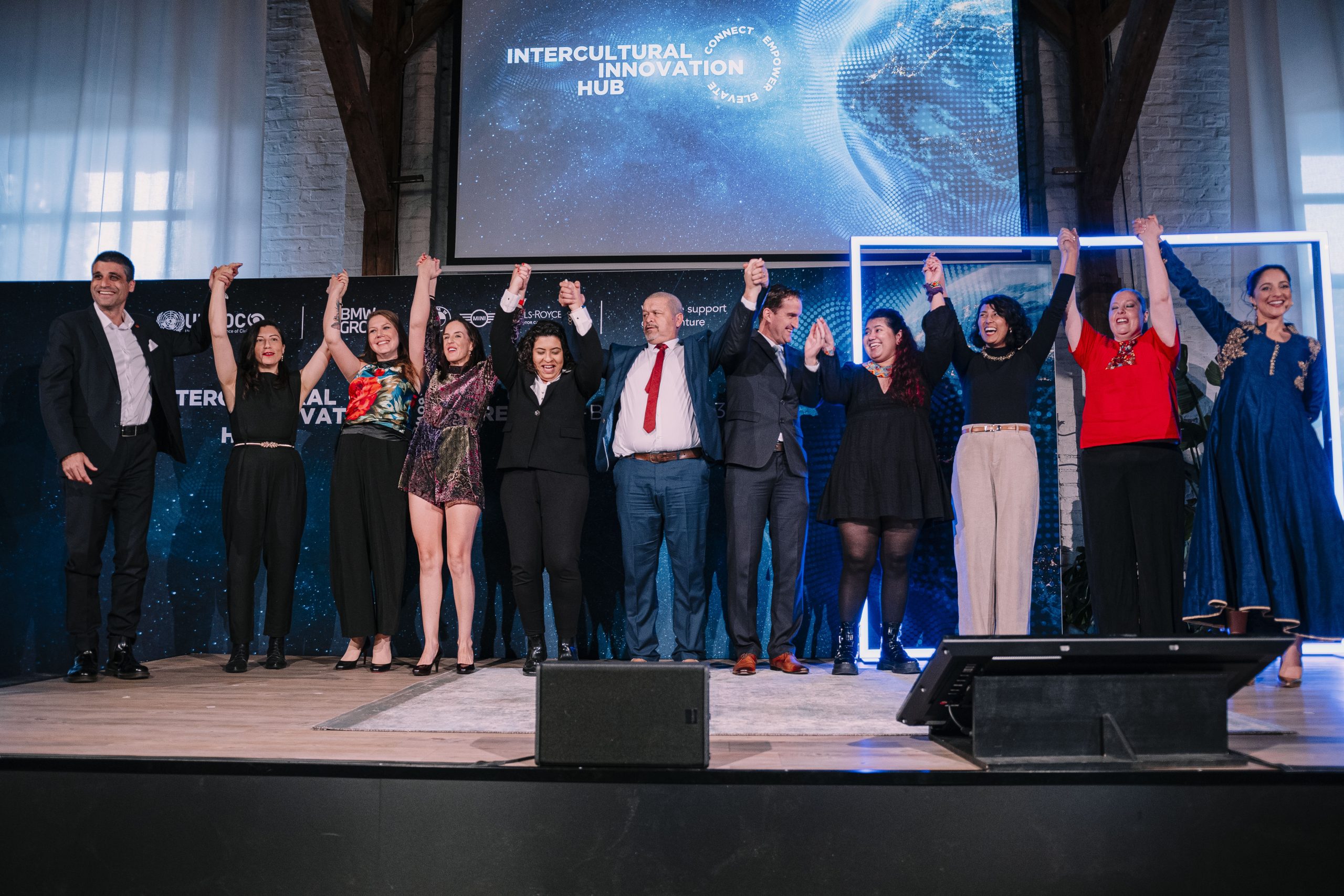 Grupo de pessoas de mãos dadas e braços para o alto, em cerimônia do Intercultural Innovation Hub. Eles estão em um palco, com um telão no fundo. 