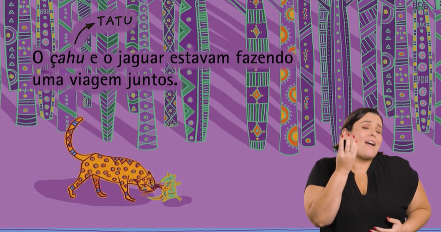 Captura de tela do livro em formato audiovisual acessível "Olhos de Jaguar". Em um fundo roxo, com totens coloridos. Lê-se "O tatu e o jaguar estavam fazendo uma viagem juntos". Abaixo do texto, estão ilustrações do jaguar e o do tatu. No canto inferior direito, está a tradutora e intérprete em Libras.