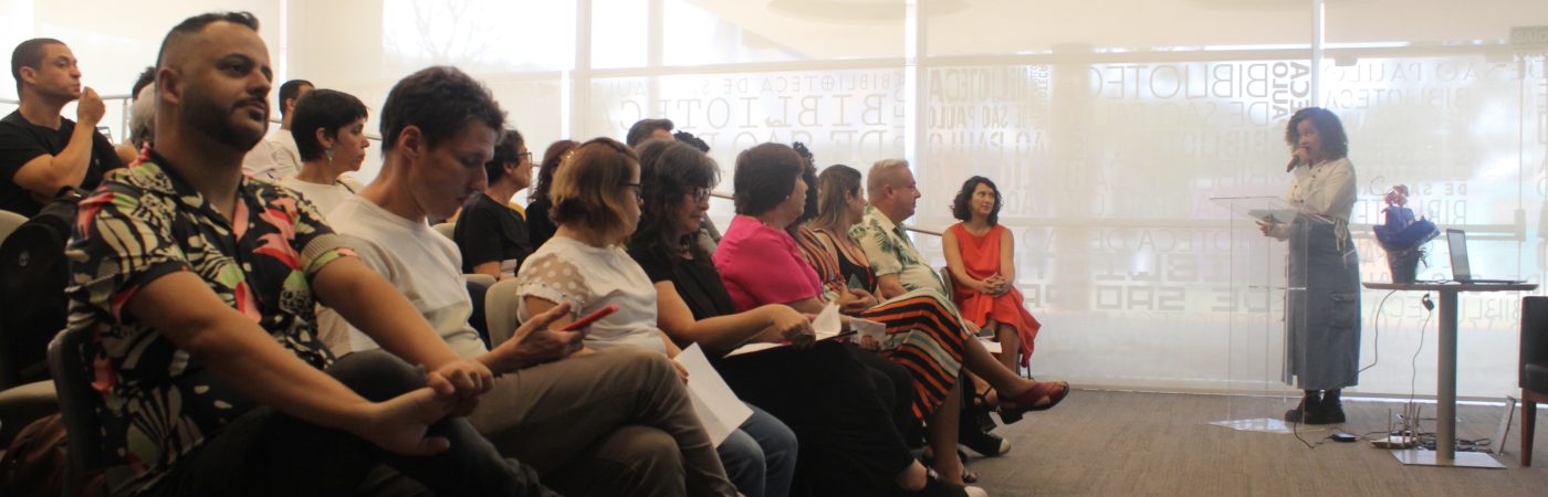 Foto colorida do auditório da Biblioteca São Paulo, que mostra as primeiras fileiras de cadeiras ocupadas pelos convidados do evento, de frente para Lara Souto Santana, que fala ao microfone, por detrás de um púlpito de acrílico.
