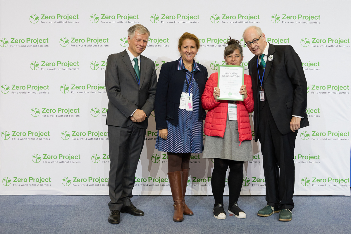 Foto colorida que mostra os representantes do Prêmio Zero Project, junto com Carla Mauch, coordenadora geral da MD, recebendo a premiação. Eles posam para a câmera, sorrindo. Ao fundo, está um painel com os logos do Zero Project, lado a lado.