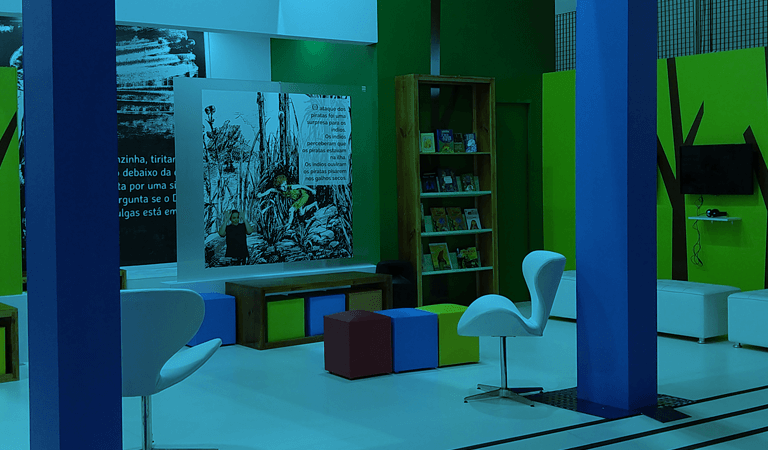 Visão do estande de livros em múltiplos formatos acessíveis na Bienal 2018. Há bancos coloridos e cadeiras brancas. Uma estante com livros e monitores de TV afixados em paredes verdes. 