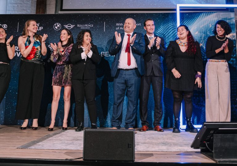 Foto de um grupo de representantes das organizações reconhecidas na cerimônia do Intercultural Innovation Hub. Eles estão sobre um palco com um telão de fundo. Eles sorriem e batem palmas.
