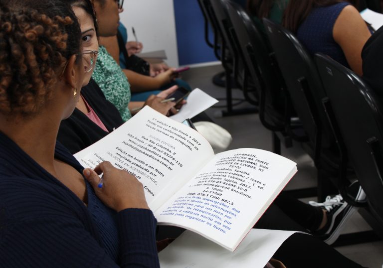 A imagem mostra uma pessoa folheando um livro em braille. O livro parece ser uma edição em braille e fonte ampliada de um livro infantil chamado "Pedro vira porco-espinho", publicado pela Jujuba Editora. Ao lado do livro, há uma ficha catalográfica com informações sobre a publicação. A pessoa que está manuseando o livro parece estar em um ambiente de sala de aula ou biblioteca, com outras pessoas ao redor.