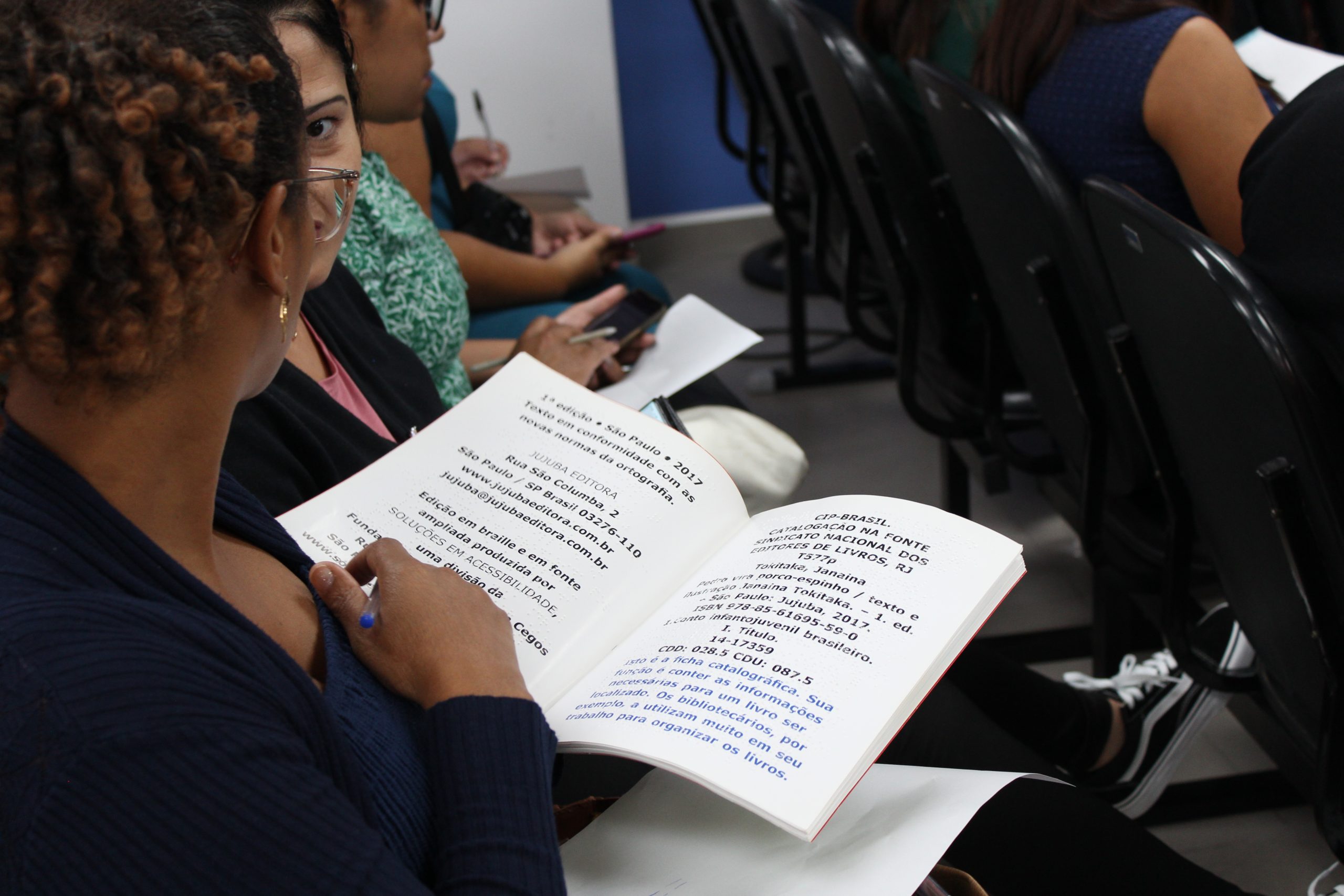 A imagem mostra uma pessoa folheando um livro em braille. O livro parece ser uma edição em braille e fonte ampliada de um livro infantil chamado "Pedro vira porco-espinho", publicado pela Jujuba Editora. Ao lado do livro, há uma ficha catalográfica com informações sobre a publicação. A pessoa que está manuseando o livro parece estar em um ambiente de sala de aula ou biblioteca, com outras pessoas ao redor.