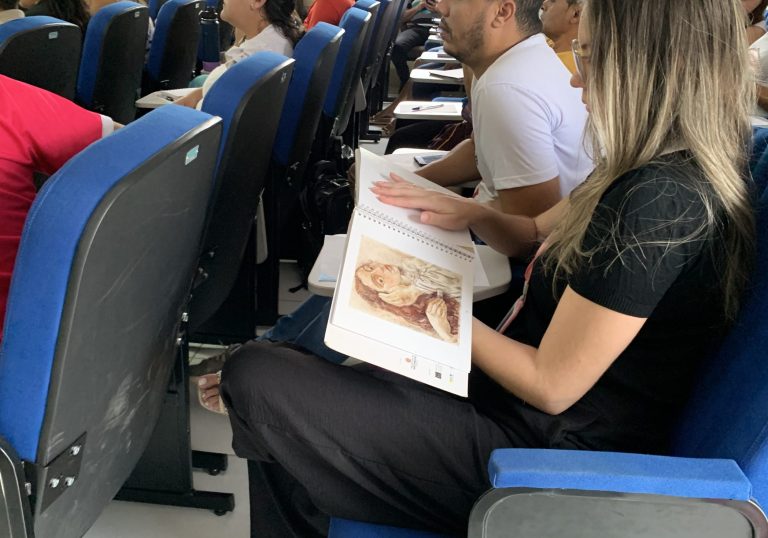 A imagem mostra uma sala de aula ou auditório lotado com diversos estudantes sentados em cadeiras azuis. Eles parecem estar envolvidos em alguma atividade ou aula, pois muitos deles estão com cadernos e materiais escolares. Na frente da sala, há um casal sentado em uma das cadeiras, aparentemente interagindo com o conteúdo que está sendo apresentado. Um deles segura uma espécie de imagem ou material impresso. O ambiente é organizado e estruturado, com paredes claras e um painel de fundo escuro, indicando tratar-se de um espaço educacional ou de palestra. A imagem transmite a sensação de uma aula ou evento em andamento, com os alunos engajados e atentos.