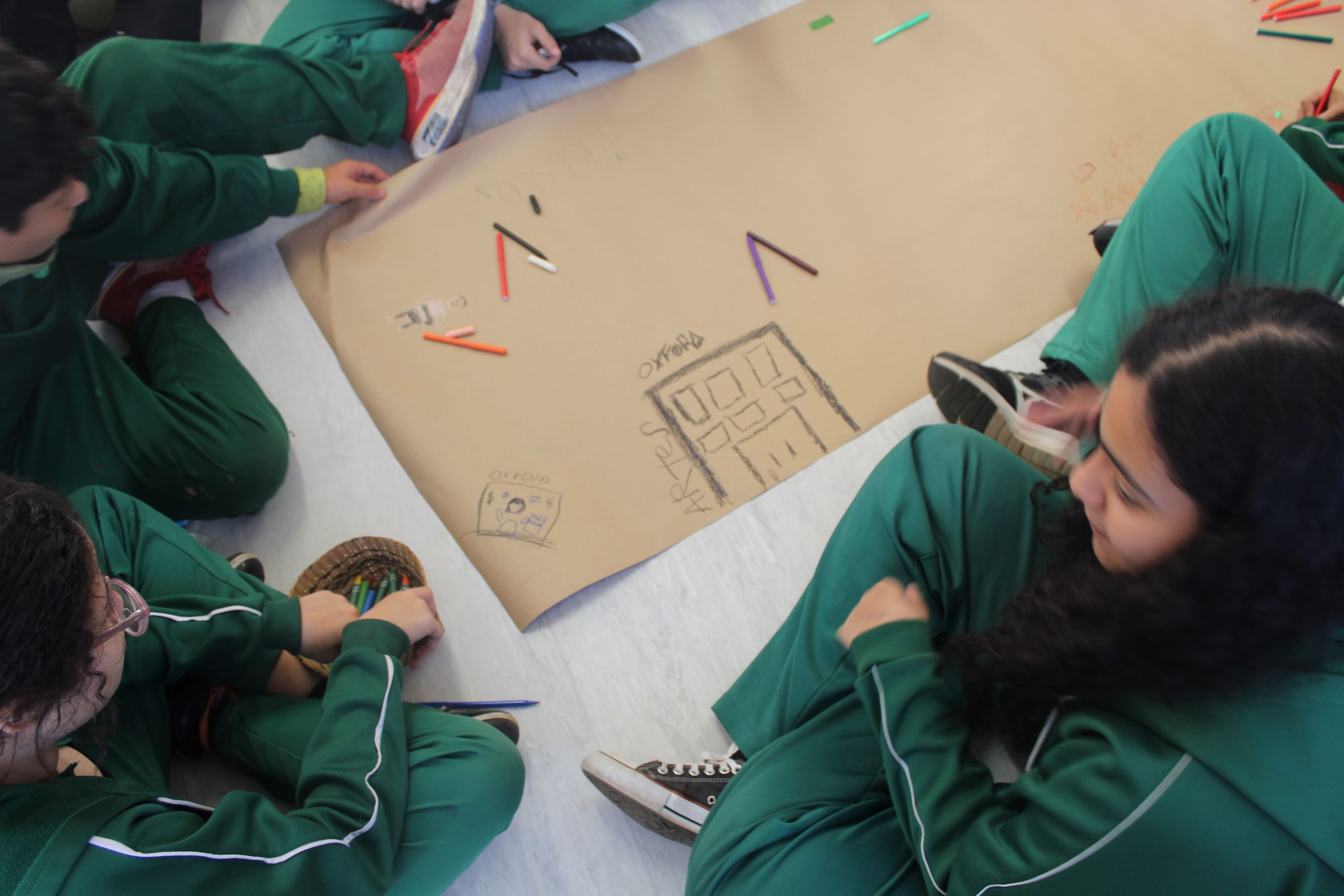 Foto de um grupo de estudantes sentados no chão, eles estão desenhando em um papelão coletivo.
